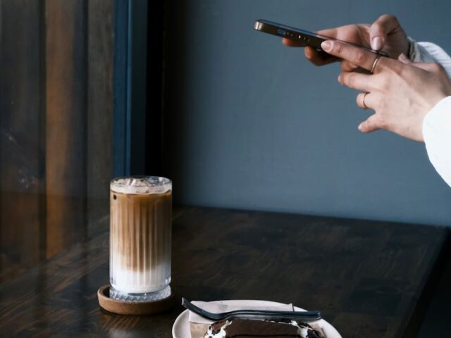 a person taking a photo of a glass with iced coffee and a dessert on a plate