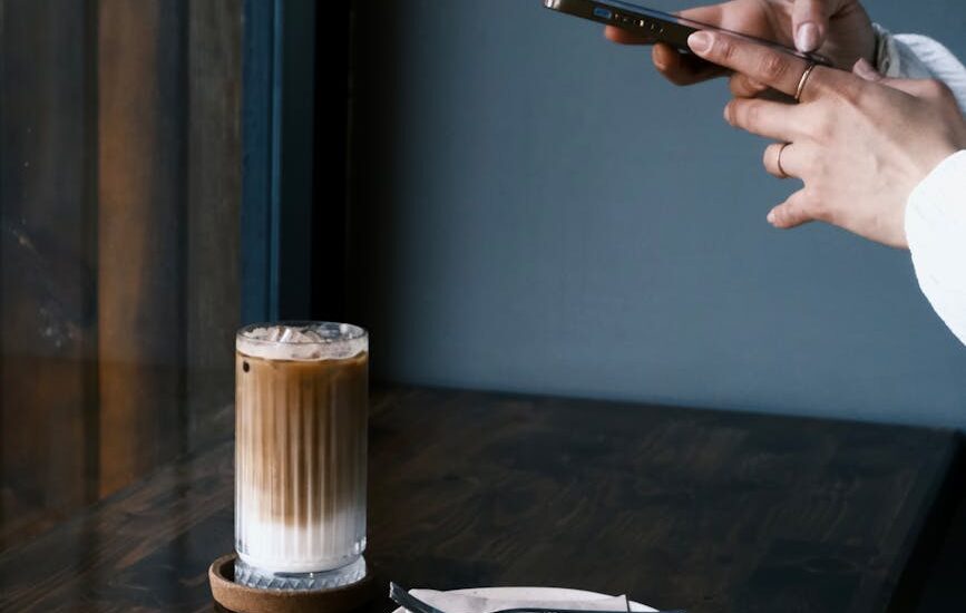 a person taking a photo of a glass with iced coffee and a dessert on a plate